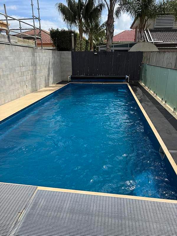 Long lap pool with a pool cover roller and glass balustrade beside a dark timber fence